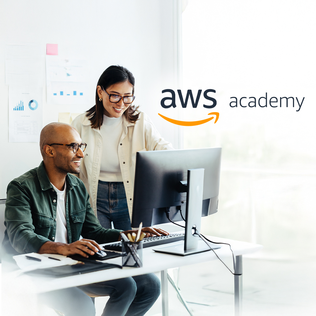 A diverse male and female duo smile while looking at a desktop computer. The woman stands behind the man, who sits at the desk and holds a mouse. The Amazon Web Services (AWS) Academy logo is in the top right corner.